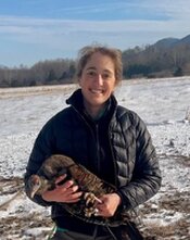 A young woman wearing a winter coat holds a bird that has been banded for identification. She stands on snowy ground with trees and partly sunny skies in the background.