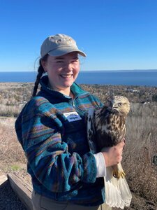 Young woman smiling, wearing a ball cap and holding an owl. The background is open ground with water on the horizon and clear blue skies.